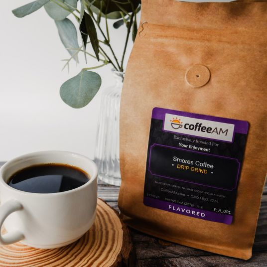 A brown kraft-paper coffee bag labeled coffeeAM Smores Coffee (Drip Grind) stands upright on a rustic wooden surface. Positioned directly to the left of the bag is a white ceramic cup filled with black coffee resting on a wooden coaster. To the left behind the cup sits a clear glass vase containing green eucalyptus leaves.