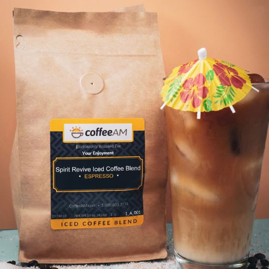 A bag of Coffee AM Spirit Revive Iced Coffee Blend Espresso stands next to a tall glass of iced coffee with a small yellow floral paper umbrella, against a peach-colored background.