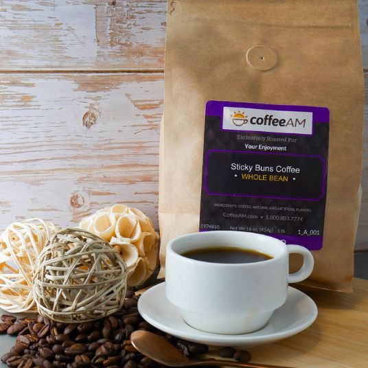 A kraft-paper bag labeled "coffeeAM Sticky Buns Coffee, WHOLE BEAN" sits on a rustic wooden background. In front of the bag is a white ceramic cup of black coffee on a matching saucer, flanked by scattered coffee beans, a wooden spoon, and decorative wicker balls.