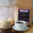 A kraft-paper bag labeled "coffeeAM Sticky Buns Coffee, WHOLE BEAN" sits on a rustic wooden background. In front of the bag is a white ceramic cup of black coffee on a matching saucer, flanked by scattered coffee beans, a wooden spoon, and decorative wicker balls.