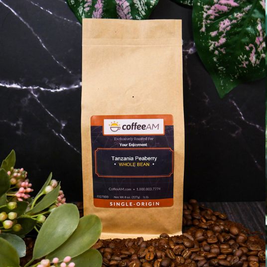 CoffeeAM Tanzania Peaberry coffee bag on a dark surface with coffee beans and greenery.