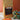 A kraft-paper bag of CoffeeAM Uganda Mount Elgon Espresso grind coffee with a dark label stands on a rustic white wooden surface against a terracotta background, with a colorful mosaic elephant figurine and a red-and-yellow woven coaster nearby.