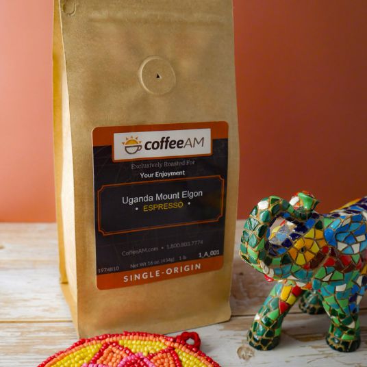 A kraft-paper bag of CoffeeAM Uganda Mount Elgon Espresso grind coffee with a dark label stands on a rustic white wooden surface against a terracotta background, with a colorful mosaic elephant figurine and a red-and-yellow woven coaster nearby.