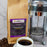A bag of coffeeAM Vienna Strudel Coffee whole bean next to a stainless steel French press and a white cup of black coffee on a marble surface with coffee beans scattered around.