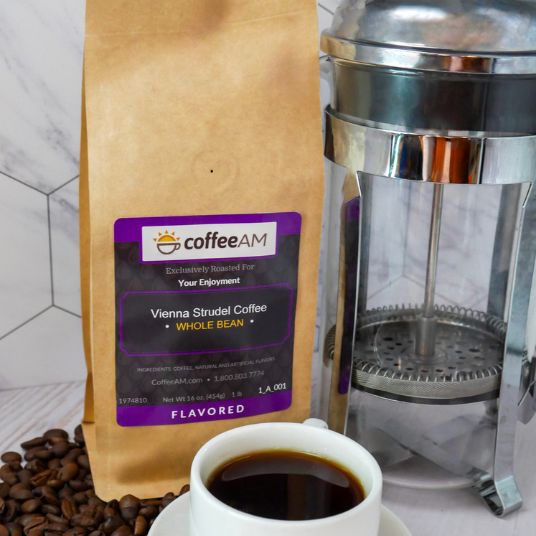 A bag of coffeeAM Vienna Strudel Coffee whole bean next to a stainless steel French press and a white cup of black coffee on a marble surface with coffee beans scattered around.