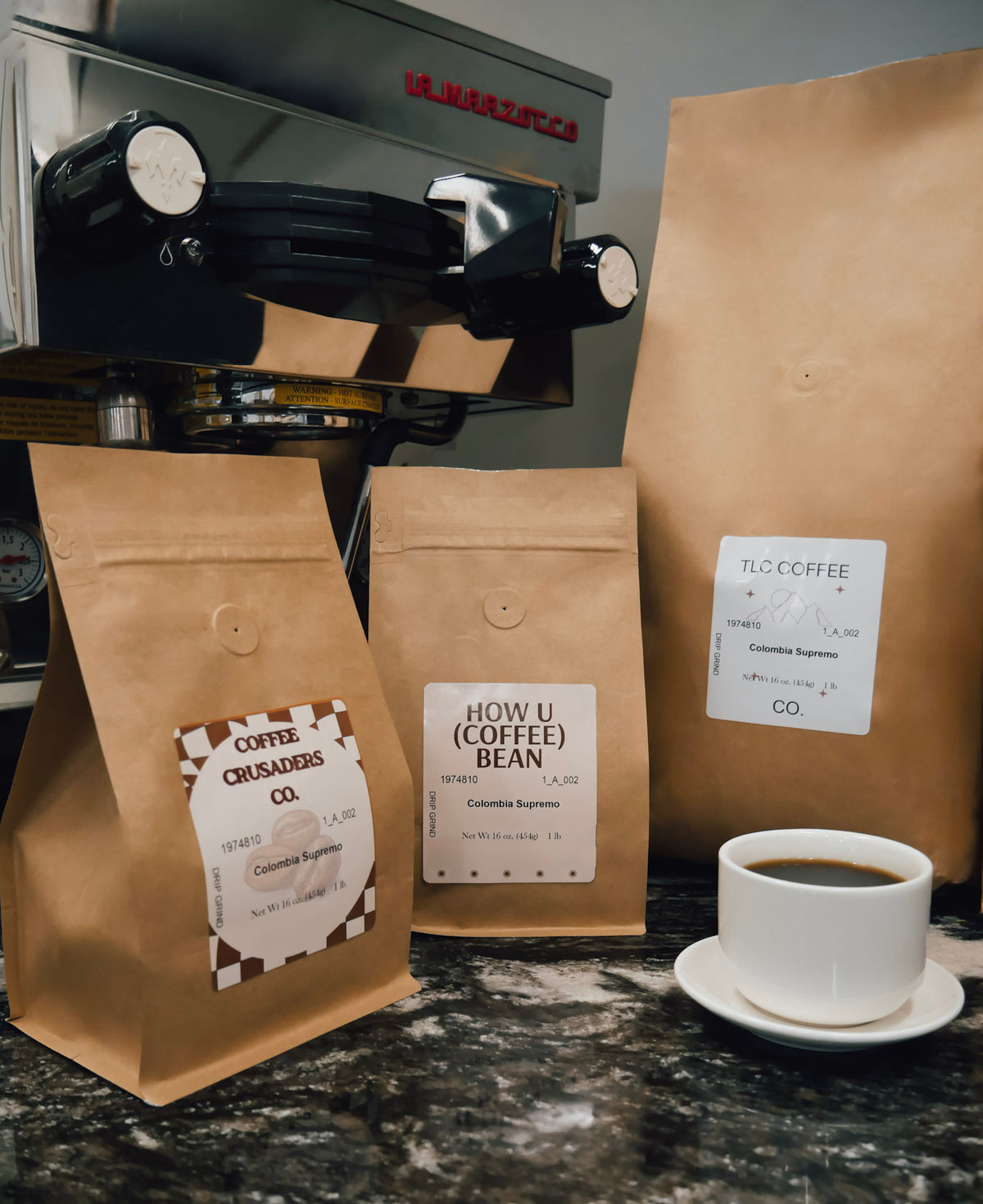 Three brown kraft coffee bags with white labels are arranged on a dark countertop in front of a coffee machine, with a white cup of espresso on a saucer to the right. Visible labels include Coffee Craze Co.
