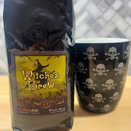 Bag of 'Witches Brew' coffee and a black mug with skull and crossbones pattern on a wooden surface.